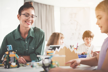 Positive teacher looking at smiling girl playing with electronic toy while situating at table