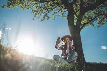 Hello. Full length of nice young woman sitting on green meadow in summer. She is full of happiness and joy holding cellphone and waving hand. Copy space in left side