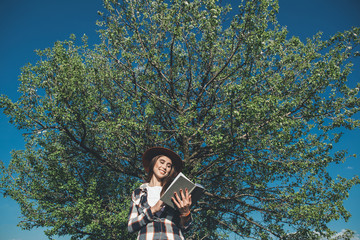 Adorable story. Low angle waist up of pretty girl standing close to green tree and admiring book with smile on face. She is pleased with spending time outside
