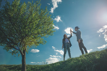 Love me like you do. Low angle full length of girl and boy standing on green hill among beautiful summer nature and touching hands. They are calm and happy with amazing blue sky on background