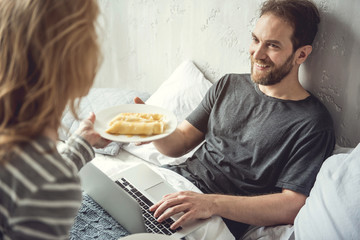 Loving woman is offering sweet pancakes to beloved partner. Happy boyfriend is sitting on cot and...