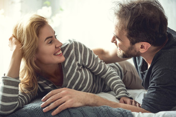 Cheerful bearded man is embracing beloved woman. She is looking at soulmate with love and joy. They are lying on couch in domestic atmosphere an enjoying themselves