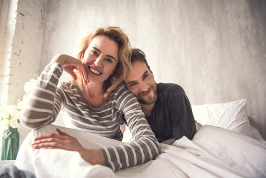 Waist up portrait of tender smiling spouse looking at camera. Man is gently putting head on woman shoulder while she is leaning to be closer to him. They are sitting on large white cot feeling happy