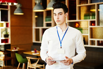 Fototapeta premium Confident young man wearing white shirt and Id card standing with tablet in coffee break room and looking at camera.