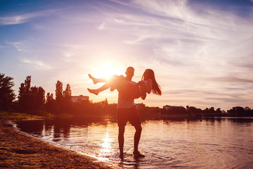 Young man holding his girlfriend in his hands. Couple having fun on summer river bank at sunset. Guys relaxing