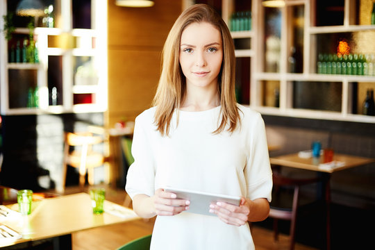 Young businesswoman in casual clothes holding tablet computer and looking confidently at camera standing in cafeteria.