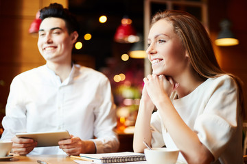 Young cheerful man and woman sitting with notepads and coffee cups at table in cafe, looking away and smiling.