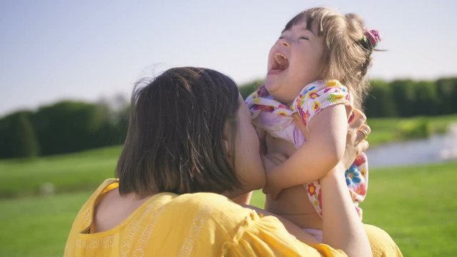 Mother and down syndrome daughter enjoying together outdoors on green grass meadow. Mom tickling little special needs girl making her laugh out of happiness. Funny family laughing together