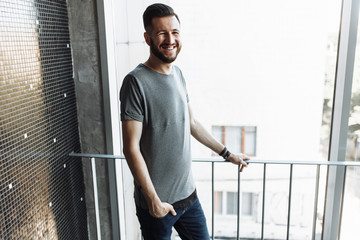 Photo of a beautiful stylish guy, hipster, dressed in a gray empty t-shirt, standing near the window, on a black wall background. Empty space for logo or design.