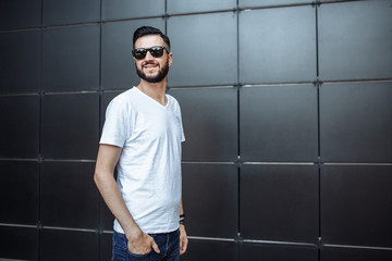 Photo of a beautiful stylish guy, a hipster with glasses, dressed in a white empty t-shirt, standing on a black wall background. Empty space for logo or design.