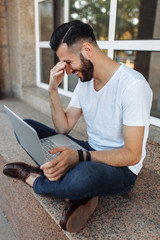 Portrait of a stylish and positive Guy, in a white t-shirt, who sits on the parapet, working with a laptop, on the street in front of a glass building, can be used for advertising, text insertion