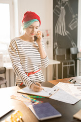 Portrait of creative female fashion designer speaking by phone and drawing sketches for custom made clothes while standing at work table in sunlit atelier workshop