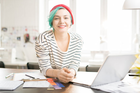 Portrait of contemporary creative young woman with mint-colored hair smiling happily looking and camera while standing at work table in sunlit design studio