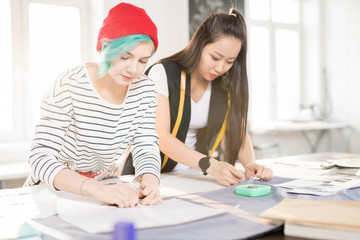 Portrait of two contemporary young women making patterns for custom made clothes on tailors table while collaborating on creative fashion design project in small atelier studio, copy space
