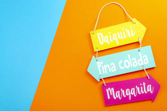 Happy hour, alcohol and summer vacation drinks concept with a wooden menu sign for daiquiri, margarita and pina colada cocktails isolated on minimalist orange and blue background with copy space - Powered by Adobe