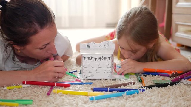 Young mother and her beautiful daughter, paint a paper house, lying on the floor at home, lifestyle, creativity, education.