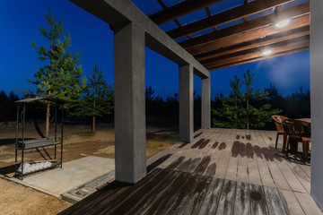 Side view of an open veranda in front of a modern forest cottage. Pine young forest under the sunset rays in the background.