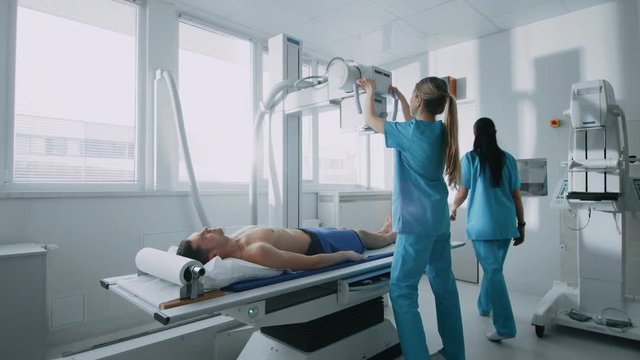 In the Hospital, Low Angle Shot of Man Lying on a Bed, Female Technician adjusts X-Ray Machine. Scanning for Fractures, Broken Limbs, Injuries, Cancer or Tumor. 