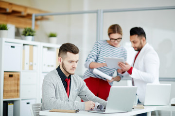Serious businessman sitting at laptop and writing in document at his workplace