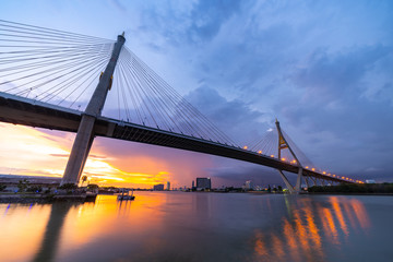 Obraz premium Bhumibol Bridge, the Industrial Ring Road Bridge with skyscraper in the night scene after sunset. Twilight sky and light reflection on smooth water, Thailand