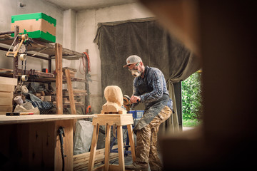 Сarpenter,small buiness owner in work clothes saw to cut out sculpture from wooden a man's head, using an angle grinder  in the workshop, around a lot of tools,wooden,furniture for work
