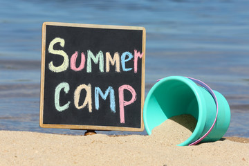 Little blackboard with text SUMMER CAMP and toy on sand near sea