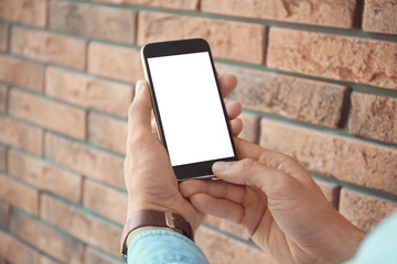 Young man holding mobile phone with blank screen in hands near brick wall