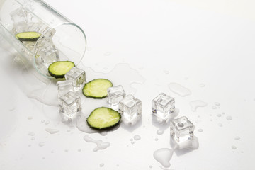 Spilled Glass with refreshing water, cucumber slices and ice cubes on a light background