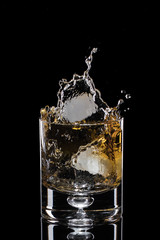 Ice falls into a glass with whiskey. The spray is flying. Black background.