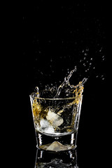 Ice falls into a glass with whiskey. The spray is flying. Black background.