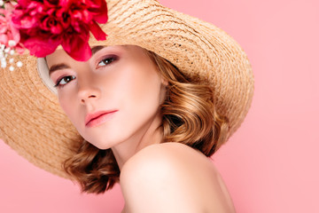 portrait of gorgeous young woman in hat with beautiful flowers looking at camera isolated on pink