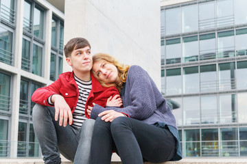 young affectionate heterosexual couple sitting on steps in front of modern building with glass facade