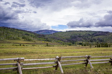 Fototapeta na wymiar Clouds Hang over the Rocky Mountains with a Wood Fence Built across a Green Meadow in the Foreground in Colorado, USA