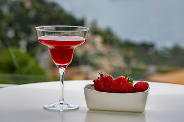 A glass with a red cocktail and strawberries on a beautiful background of mountains and the sea. Dusk. Toned.