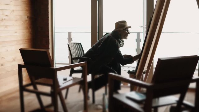 Old artist is drawing in light room. He is sitting on a chair, holding brush in hands and touching by it over canvas on easel