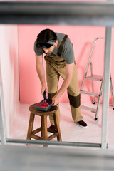 man in working overall and headband dipping paint roller into roller tray on chair