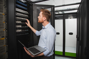 Processing system. Focused IT technician examining server closet while holding laptop