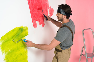 side view of smiling man in working overall painting wall by two paint rollers