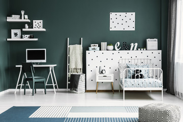 White metal twin bed by a polka dot wall with decorations and a wooden desk with computer screen in a dark green room interior for a teenager