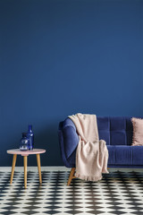 Chessboard floor, sofa with a blanket and stool with glass decorations on an empty navy blue wall