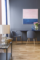 Real photo of a spacious dining room with a blue and pink painting on a grey wall, above a table and two chairs and with a table with lamp in front of the photo