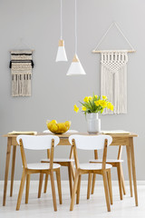White chairs at wooden table with yellow flowers in grey dining room interior with lamps. Real photo