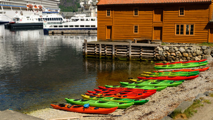 Fototapeta na wymiar Colorful canoes along the beach in front of a brown house. Ferries and cruise ship in the background. Location Eidfjord in Hordaland, Norway.
