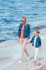 happy stylish mother and daughter holding hands and walking on sea stylish