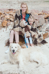 attractive mother and daughter in blanket sitting with dog on shore
