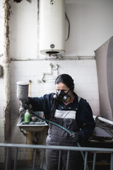 Strong and worthy woman doing hard job. She using industrial spray compressor for painting some metal products.