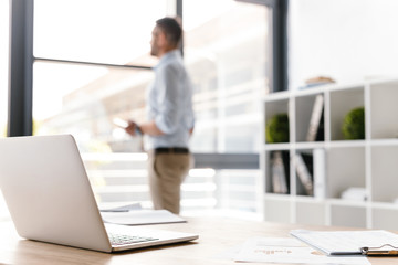 Photo of workplace with open white laptop lying on table, while defocused business man standing and looking through big window on background