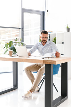Full length image of caucasian unshaved businessman 30s in white shirt working in office on laptop, and looking at clipboard with paper documents
