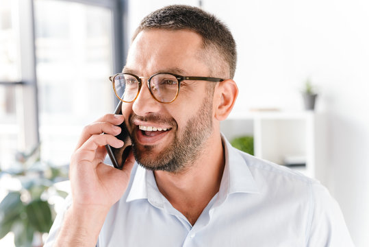 Image closeup of bearded man in white shirt smiling and talking on black smartphone about business, while working in office