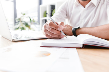 Cropped image of adult business man in white shirt sitting at table and writing down information...